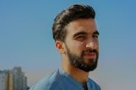 A close-up outdoor portrait of a young man with facial hair, exuding confidence under a clear sky.